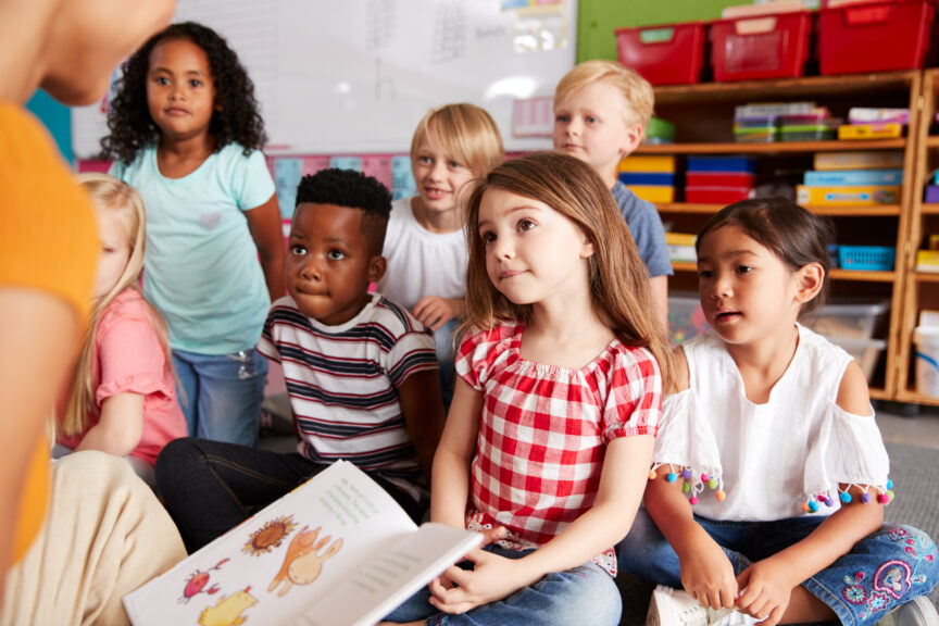 Group Of Elementary School Students Sitting On Floor Listening To Teacher Read