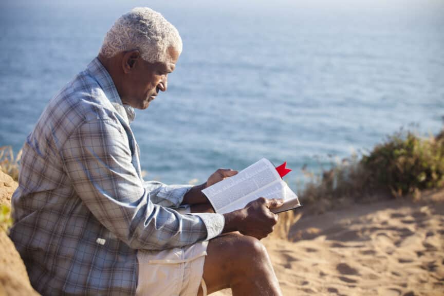 African American Man Reading Bible On Beach