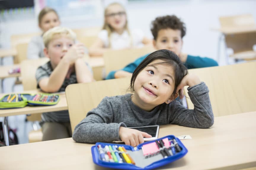 Portrait of schoolgirl looking up towards teacher while sitting in classroom with classmates