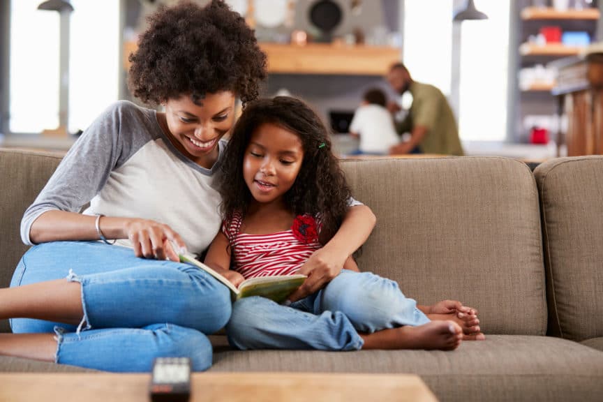 Mother And Daughter Sit On Sofa In Lounge Reading Book Together