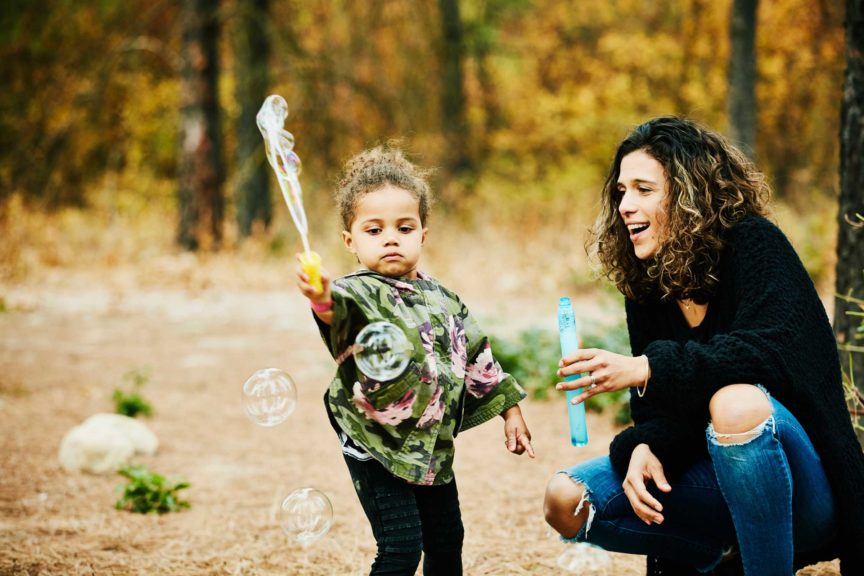 Smiling mother watching young daughter play with bubbles outside