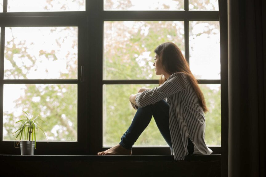 Thoughtful girl sitting on sill embracing knees looking at window