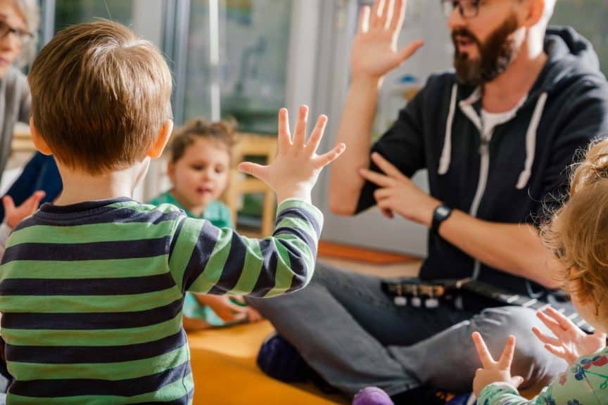 children with hands raised sitting on floor with teacher 