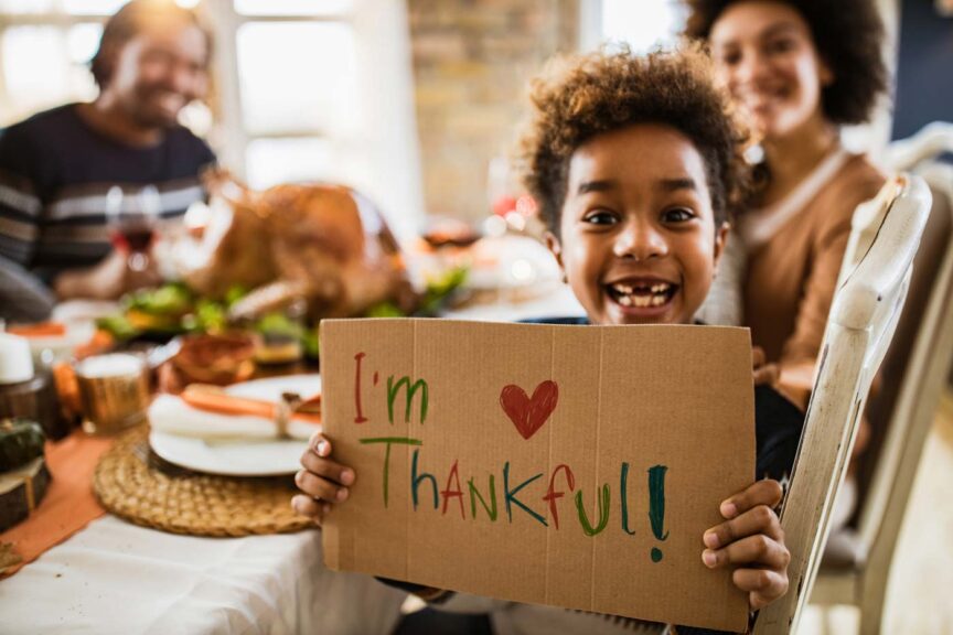 girl holding 'I'm thankful' sign and looking at camera during Thanksgiving meal with her parents