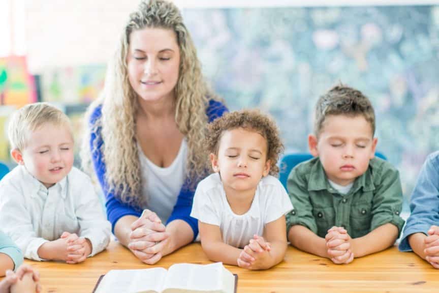 Kids and teacher are sitting at a table and praying with their teacher.