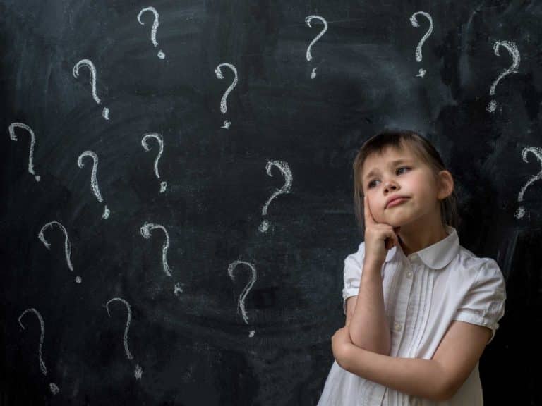 Thoughtful Cute Girl Looking Away While Standing Against Blackboard with multiple question marks written on it