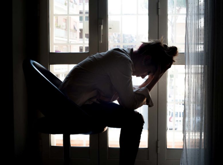 Tensed Woman With Head In Hands Sitting On Chair By Window At Home